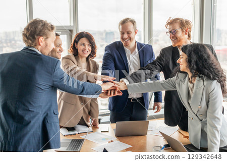 A diverse business team of six people stand around a table with laptops and paperwork in a modern office. They have their hands stacked in the center, smiling and celebrating a shared success. 129342648