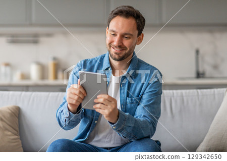 A man in a blue shirt is sitting on a white couch in a living room. He is smiling as he looks at a tablet in his hands. 129342650