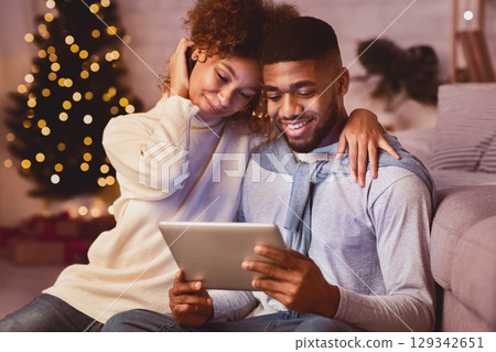 Joyful black couple using digital tablet against Christmas tree, enjoying winter evening at home 129342651
