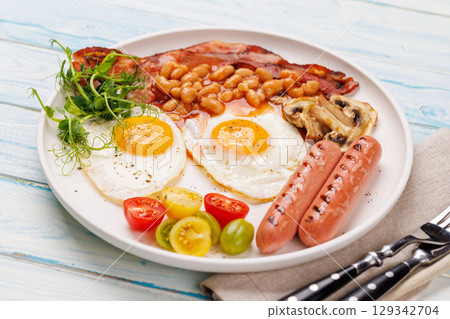 A classic breakfast plate featuring sunny-side-up eggs, crispy bacon, sausages, and fresh tomatoes 129342704