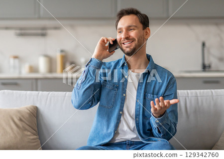 A man with a beard is sitting on a white couch in his home while talking on his phone. He is wearing a blue denim shirt and jeans. He is smiling and gesturing with his hand as he speaks. 129342760