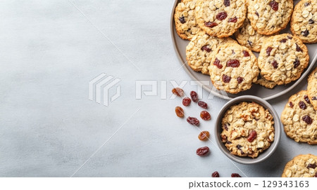 A plate filled with freshly baked oatmeal cookies scattered with raisins, accompanied by a bowl of cookies on a light gray surface. 129343163