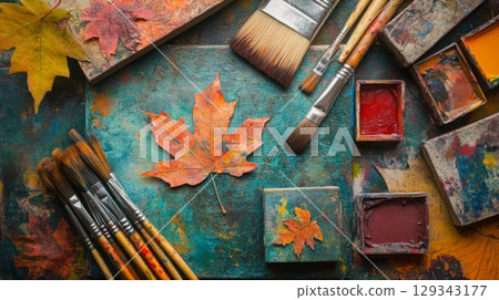 An assortment of paintbrushes and vibrant paint squares are arranged alongside autumn leaves on a textured surface, creating an inspiring atmosphere for artistic expression. 129343177