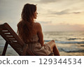 A woman sitting alone in a wooden deck chair facing a calm ocean horizon at sunset 129344534