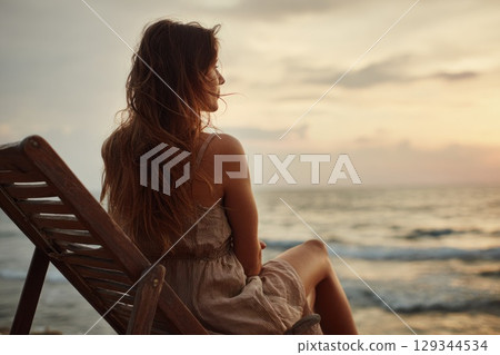 A woman sitting alone in a wooden deck chair facing a calm ocean horizon at sunset 129344534