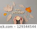 Hello october text and woman hands holding a cup of coffe. Top view, autumn leaves on grey background 129344544