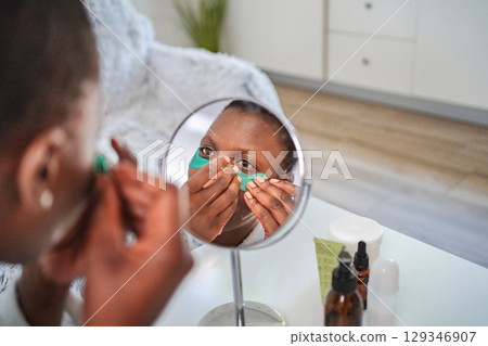 Young woman applying hydrogel eye patches in front of a mirror 129346907