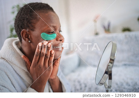 Smiling woman applying hydrogel eye patches looking in the mirror 129346910