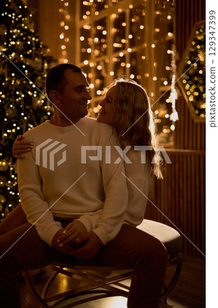 A loving couple in white sweaters shares an intimate moment, embracing in a cozy, festive room with A loving couple in white sweaters shares an intimate moment, embracing in a cozy, festive room with 129347399