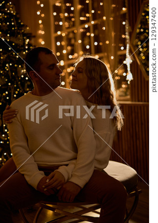 A loving couple in white sweaters shares an intimate moment, embracing in a cozy, festive room with 129347400