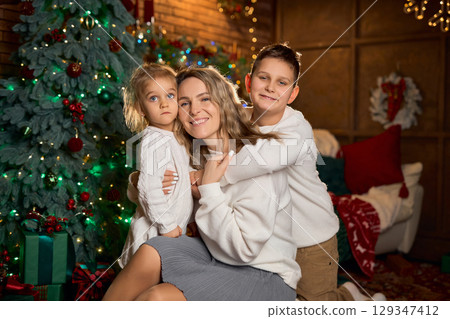 smiling mother and her two children in white sweaters hug closely, celebrating in a cozy, festive smiling mother and her two children in white sweaters hug closely, celebrating in a cozy, festive 129347412