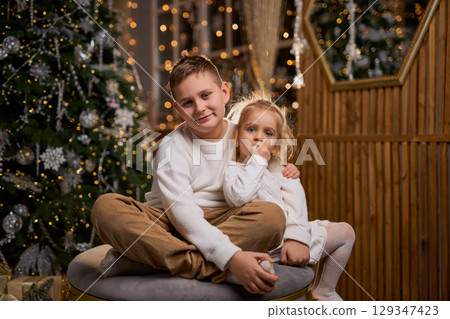A boy and his little sister, both in white sweaters, smile happily while sitting together in a cozy 129347423