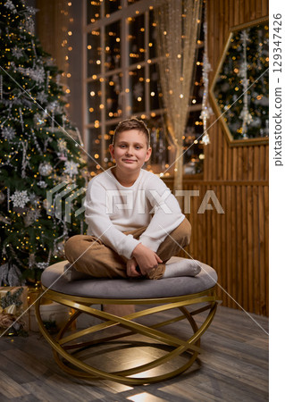 A smiling boy in a white sweater sits thoughtfully for a holiday photo, with a decorated Christmas A smiling boy in a white sweater sits thoughtfully for a holiday photo, with a decorated Christmas 129347426