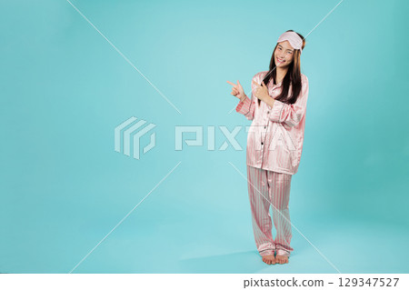 Happy young asian woman in pajamas sleep eye mask rests pointing index finger at blank area, relaxing at home she is calmly pointing left in sleepwear, full body isolated studio blue background Happy young asian woman in pajamas sleep eye mask rests pointing index finger at blank area, relaxing at home she is calmly pointing left in sleepwear, full body isolated studio blue background 129347527