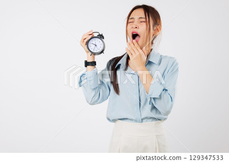 Asian woman sleep deprived and tired yawns holding time clock in frustration due to overtime, needs to wake up but is sleepy, and wants to ignore alarm clock to rest, isolated studio white background 129347533