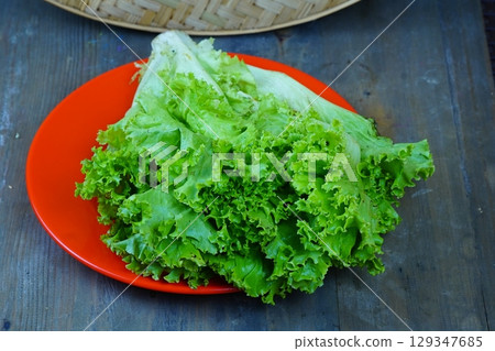 A vibrant pile of fresh, green lettuce with ruffled leaves sits on a bright red plate against a rustic wooden background. A vibrant pile of fresh, green lettuce with ruffled leaves sits on a bright red plate against a rustic wooden background. 129347685