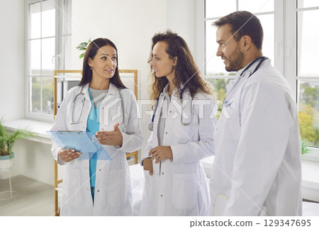 Happy team of young doctors in white uniform standing in hospital or clinic on meeting. 129347695