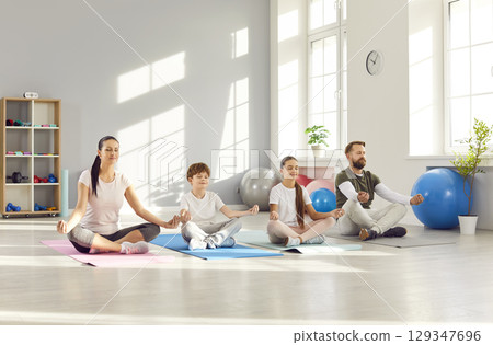 Family sitting on the floor on mat and practicing yoga in gym in Lotus pose together. 129347696