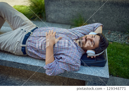 Relaxed man with headphones lying outdoors on bench 129348771