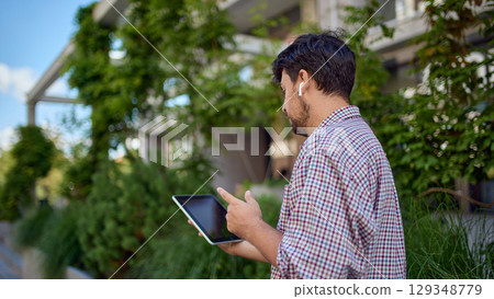Back view of businessman standing outdoors working on tablet with wireless earphones Back view of businessman standing outdoors working on tablet with wireless earphones 129348779