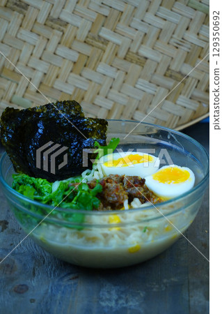 A vibrant bowl of ramen featuring noodles, savory ground meat, a soft-boiled egg, and fresh greens, topped with crispy seaweed. 129350692