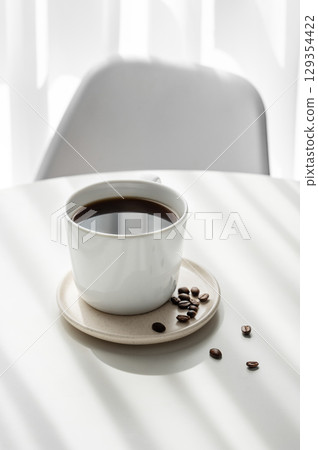 Freshly brewed espresso coffee in a cup on a white table near window with chair, coffee beans 129354422