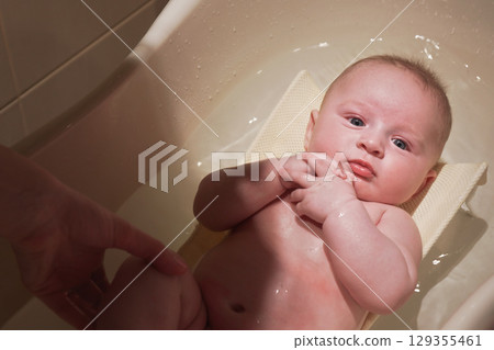 Infant baby boy washed in small bath tub, view from above, mother hand touching his leg Infant baby boy washed in small bath tub, view from above, mother hand touching his leg 129355461