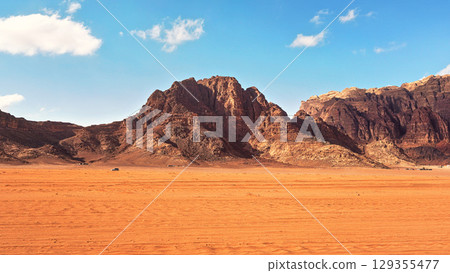 Flat red desert with large mountains in distance, blue sky above, some off road vehicles in background, typical scenery at Wadi Rum, Jordan Flat red desert with large mountains in distance, blue sky above, some off road vehicles in background, typical scenery at Wadi Rum, Jordan 129355477