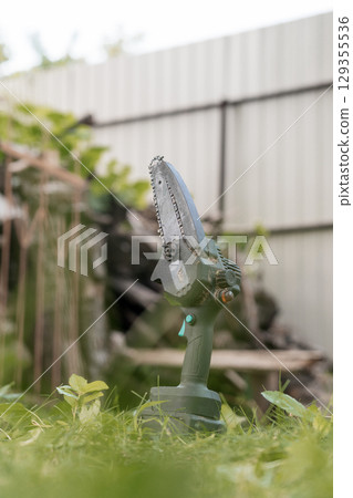 Electric One-Hand Chainsaw Cutting Tree Branch Outdoors, Green Grass, Metal Fence Background, Garden Tool, Close-Up Shot, Realistic Style Electric One-Hand Chainsaw Cutting Tree Branch Outdoors, Green Grass, Metal Fence Background, Garden Tool, Close-Up Shot, Realistic Style 129355536