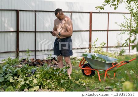 Shirtless Man Gardening Outdoors with Wheelbarrow, Brown Shorts, Green Foliage, Metal Fence, Sunny Day, Back Yard, Natural Light Photography 129355555