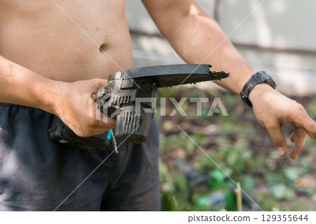 Bare-Chested Man Holding Black Hedge Trimmer Outdoors, Waist Up Shot, Garden Background, Green Foliage, Summer Day, Utility Tool, Dark Shorts. 129355644