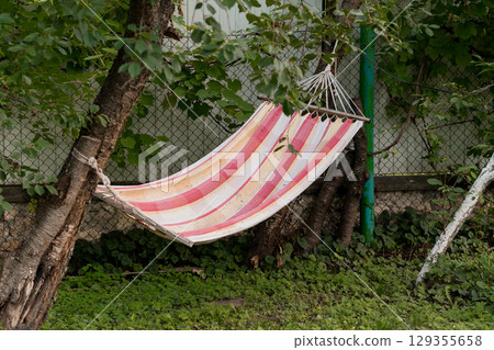 Peaceful Backyard Relaxation: Pink Striped Hammock Between Trees, Lush Green Grass, Summer Outdoor Lounging Scene with Garden Fence. 129355658