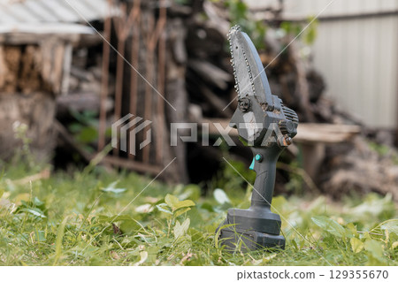 Electric Cordless Handheld Chainsaw Cutting Small Branches Outdoors, Green Grass, Rustic Wood Pile, Garden Tool, Power Equipment, Sharp Blade Electric Cordless Handheld Chainsaw Cutting Small Branches Outdoors, Green Grass, Rustic Wood Pile, Garden Tool, Power Equipment, Sharp Blade 129355670