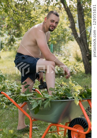 Muscular Man Gardening Bare-Chested with Wheelbarrow, Orange Tools, Green Foliage, Summer Landscape, Natural Light, Rustic Outdoor Scene. 129355831