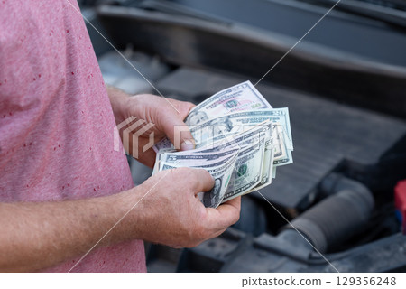 Holding US dollar cash while inspecting a vehicle under the hood in a parking lot. Person counts cash dollars while focused on the engine of a car, pondering next steps in car repairs 129356248