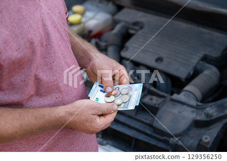 Person counting euro money while standing near open car hood in daylight. Individual checks cash and coins while leaning over an open car hood, counting car repair expenses Person counting euro money while standing near open car hood in daylight. Individual checks cash and coins while leaning over an open car hood, counting car repair expenses 129356250