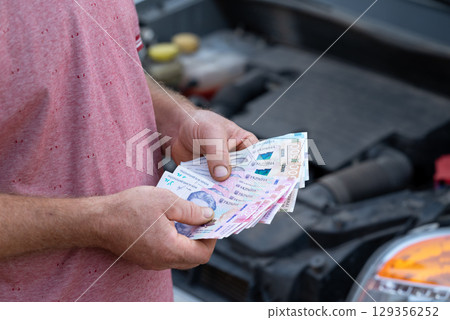 Holding Ukrainian hryvnia cash near open hood of a car. Person counts hryvnia cash near broken down car, preparing money for service payment 129356252