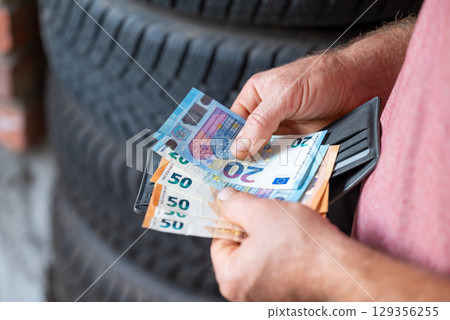Person counting euro money near tire workshop, preparing cash to pay for service. Individual is organizing Euro banknotes while standing close to tires in a workshop 129356255