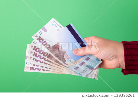 Using cash or non-cash money for payments. Hand of a female with cash hryvnia banknotes and plastic bank card on green background Using cash or non-cash money for payments. Hand of a female with cash hryvnia banknotes and plastic bank card on green background 129356261