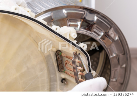 Repairing washing machine in service centre. Professional service worker taking off old broken water heater from washer to change for a new one 129356274