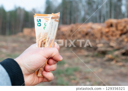 Holding euro money against cut forest. Man holding euro banknotes in front of stacked logs, representing purchase of natural resources and forest exploitation 129356321