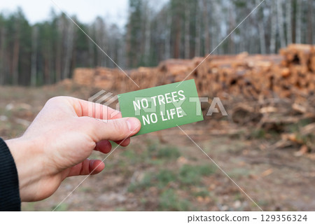 No trees no life message with deforestation background. Hand holding "No trees no life" paper near stacked logs in deforested woodland, emphasizing environmental protection and tree importance No trees no life message with deforestation background. Hand holding "No trees no life" paper near stacked logs in deforested woodland, emphasizing environmental protection and tree importance 129356324