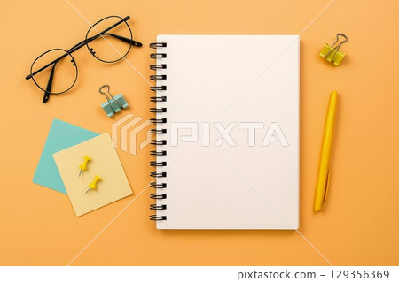 Notebook and stationery on orange background. Blank spiral notebook with pen, glasses, push pins and binder clips 129356369