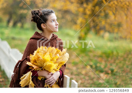 Thoughtful Girl Holding Autumn Leaves in Park. Girl wearing a brown poncho holding yellow autumn leaves while standing in a park with fallen foliage and green grass Thoughtful Girl Holding Autumn Leaves in Park. Girl wearing a brown poncho holding yellow autumn leaves while standing in a park with fallen foliage and green grass 129356849