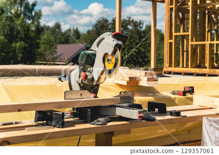 Circular saw resting on wooden plank at construction site with timber frame house being built Circular saw resting on wooden plank at construction site with timber frame house being built 129357061
