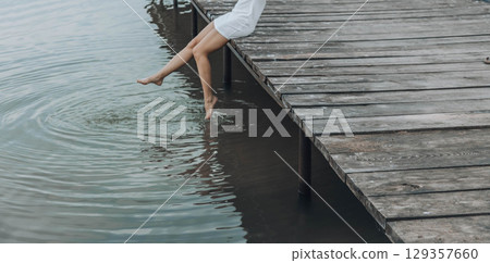 unrecognizable womans legs in a white dress, sitting on the edge of wooden pier, dipping her feet into calm water with a light splash, relaxation, travel unrecognizable womans legs in a white dress, sitting on the edge of wooden pier, dipping her feet into calm water with a light splash, relaxation, travel 129357660