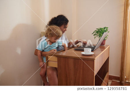 Woman Working Remotely While Caring for Her Child in a Cozy Home Space Woman Working Remotely While Caring for Her Child in a Cozy Home Space 129358781