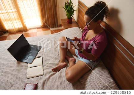 Woman Working Remotely on Laptop and Smartphone in Cozy Bedroom Woman Working Remotely on Laptop and Smartphone in Cozy Bedroom 129358810