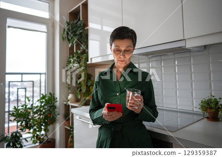 Serious woman staring at smartphone screen with water glass in kitchen, distress. Bad news stress Serious woman staring at smartphone screen with water glass in kitchen, distress. Bad news stress 129359387