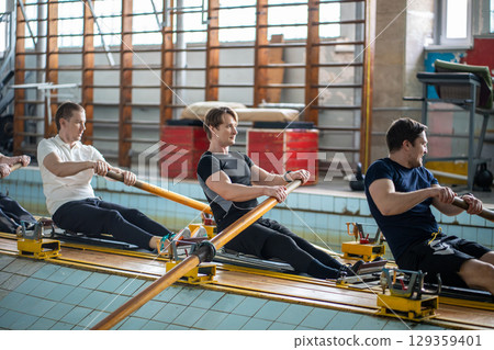 Concentrated tired athletes stroking on rowing simulator. Synchronization, water aquatic sports Concentrated tired athletes stroking on rowing simulator. Synchronization, water aquatic sports 129359401